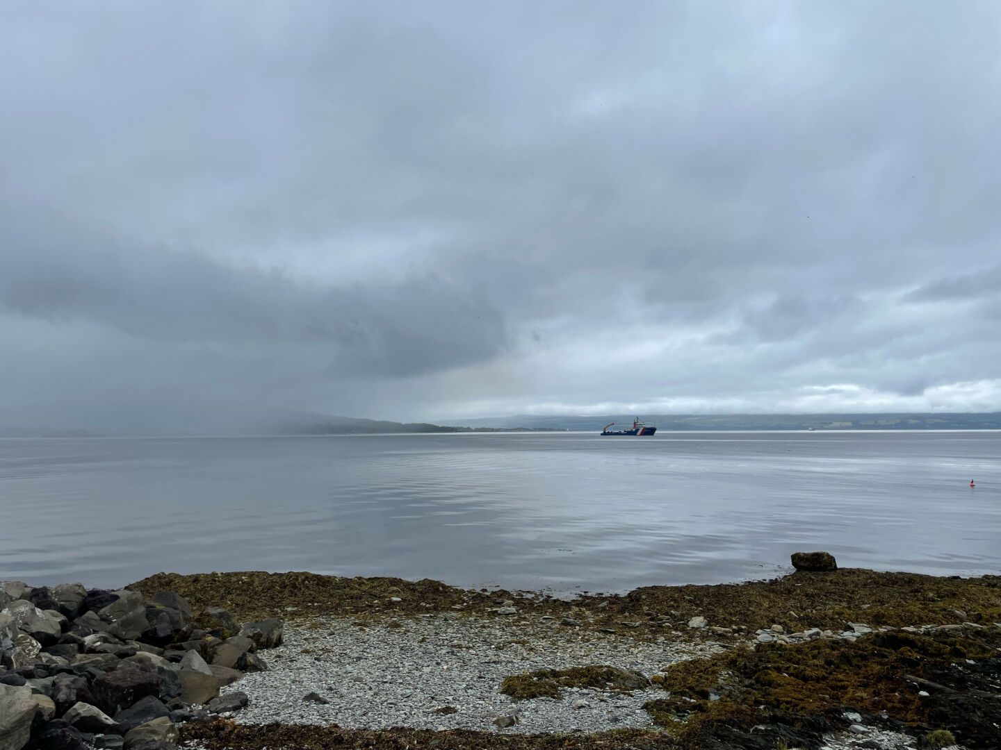 A blue ship in a bay with rain approaching from the left of the frame, a pebble beach in the foreground.