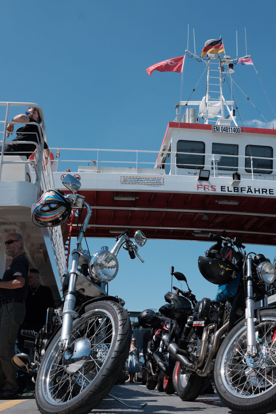 Two lines of motorcycles aboard an Elbfähre ferry. Sun beats down and flags fly.