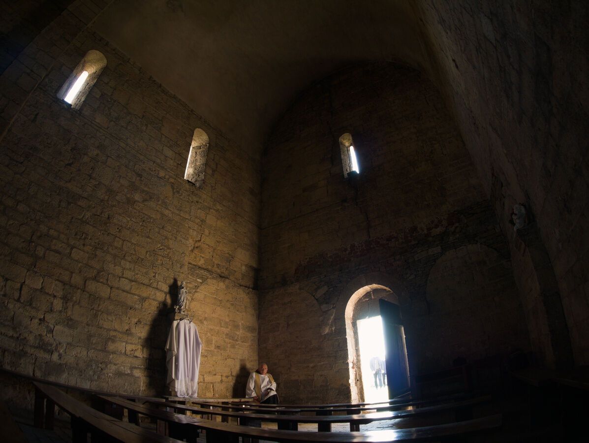 Dark interior of small Romanesque church. The church is empty, save for a priest sitting in a corner, looking towards camera. Walls and benches seem to bend around the priest (the lens used was fishey-ish).