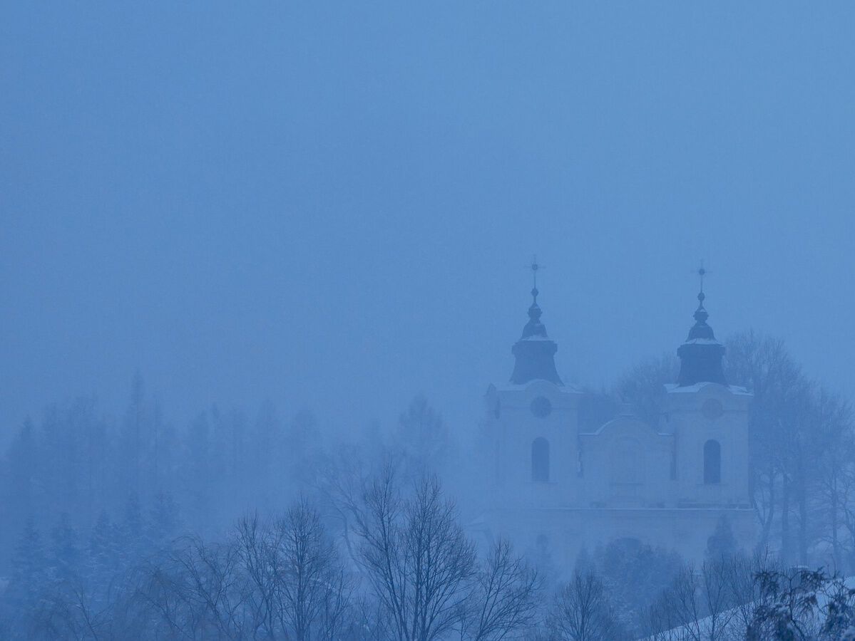 Winter scene. Evening, snow, white church on a hill, some trees. Late evening light colours everything in misty blue.