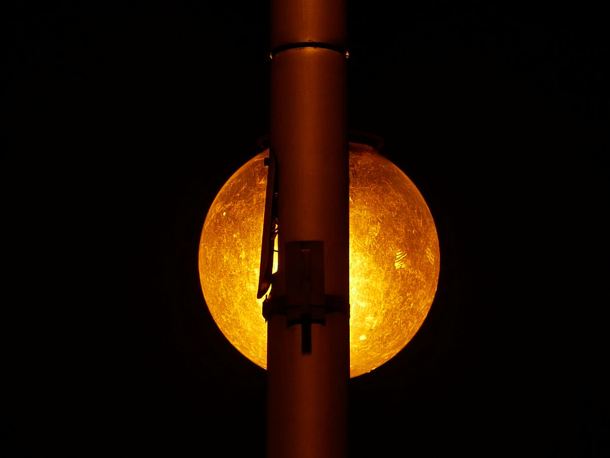 In the background — yellow-reddish glowing orb, spherical, but with heavily, irregularly textured surface. Partially obscured by long metal cylinder, crossing frame vertically. On one side of cylinder there's a kind of bay door, slightly open — the sphere is just behind this section, brigh light shines between door and the cylinder.