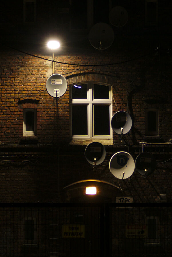 Night. Brick house wall lit by single lantern. One centrally placed window and few smaller ones, all dark and empty. Six satellite dishes of various colours and sizes, apparently placed in similar plane to the lantern, so lantern's light creates crescent-shapes inside the dishes.