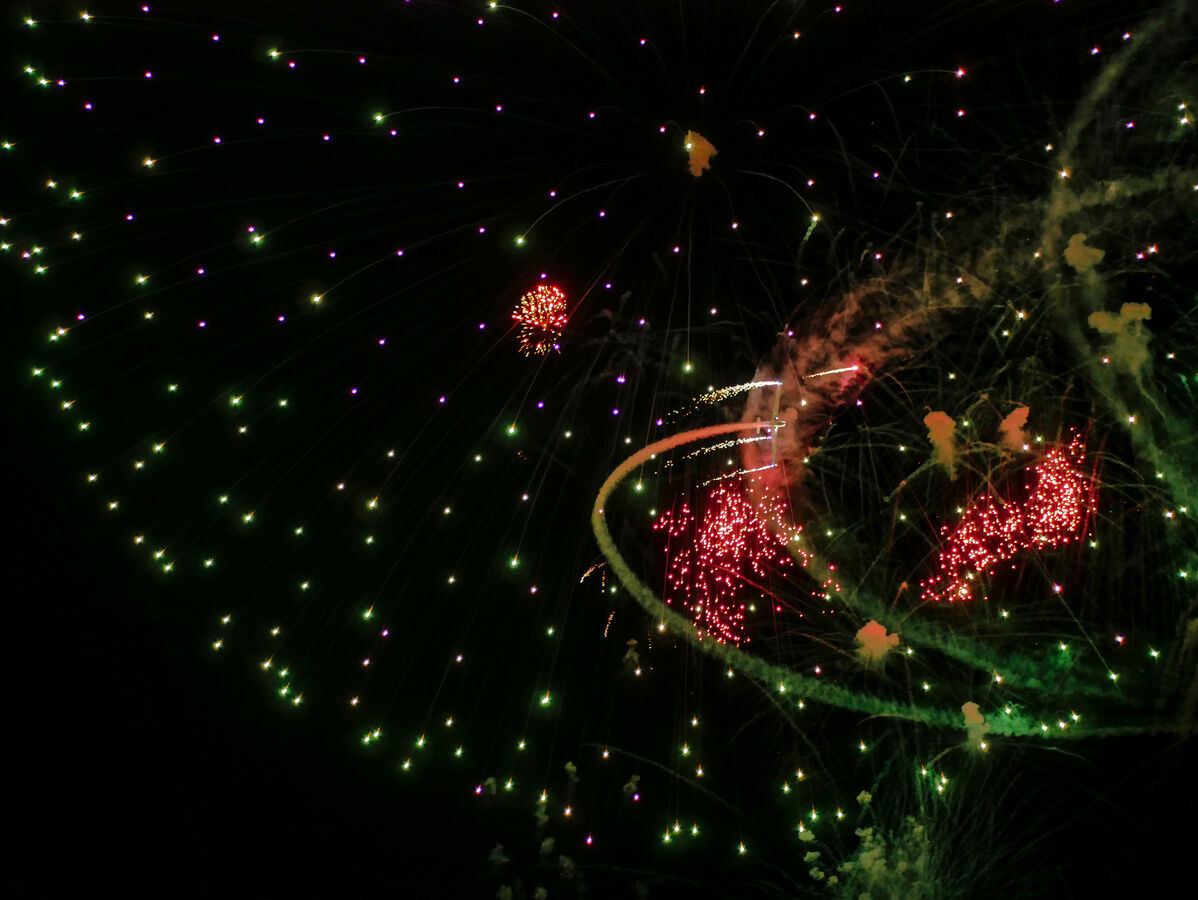 Dark night sky, fireworks. A white motoglider flies thru the fireworks, leaving smoke trail.