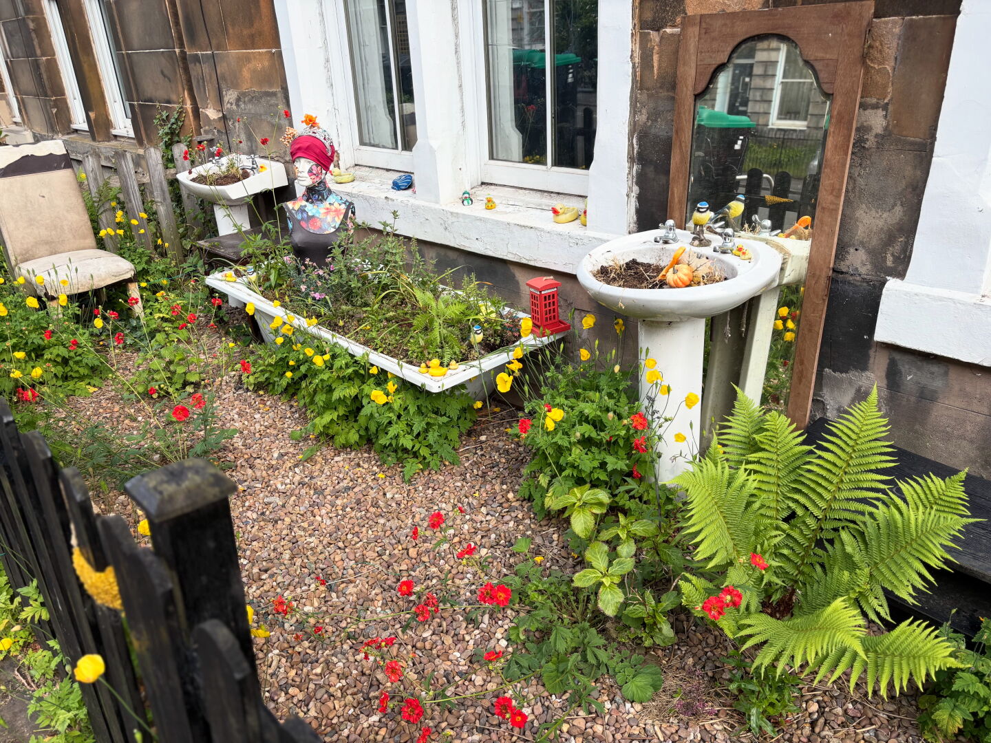 A tailor's dummy (female) seated in an old bath filled with earth, 2 washbasins, one in front of a mirror, flowers, a fern, a toy great tit, and a toy red 'phone box can all be seen in the photo.