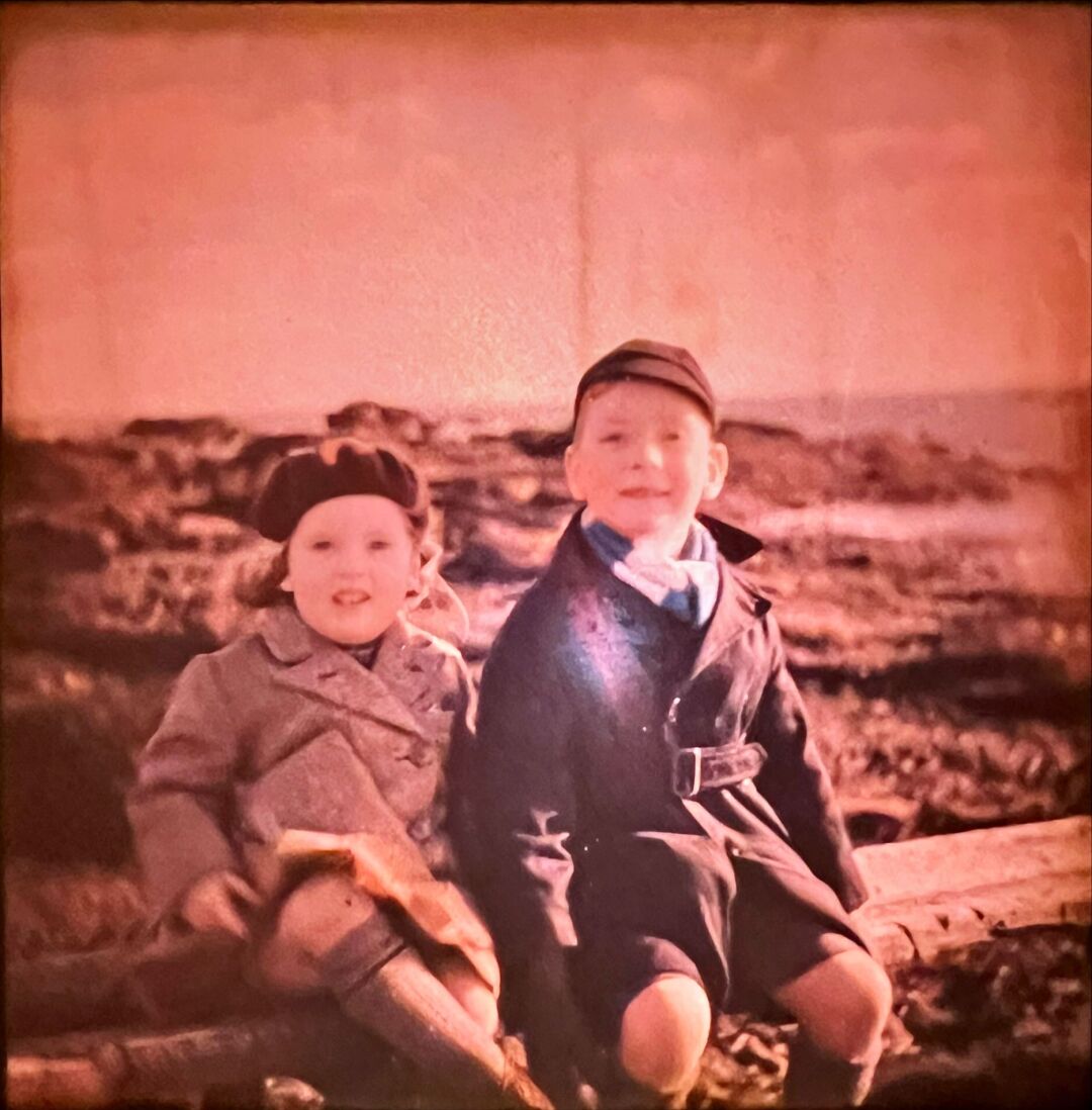 Two children are sitting on a wooden rail in the foreground, in the background there are rocks and the sea. The children are dressed in 1930s styles.