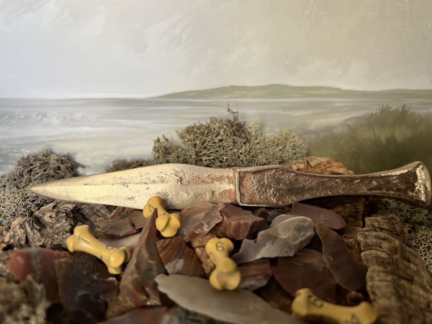 A cast bronze knife sits behind a bed of knapped flint flakes, with 4 yellowed, bone-shaped d6. The background is a landscape painting by Jon Hodgson.