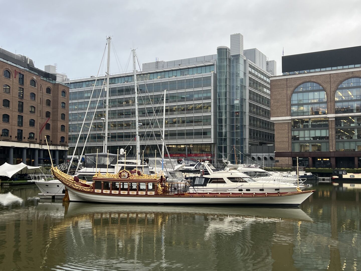 The royal barge, Gloriana, at St Katherine’s Docks. Its hull is white, and the upper parts red with many swirly gold embellishments. Behind it, there are more modern looking boats, and a wall of office buildings.