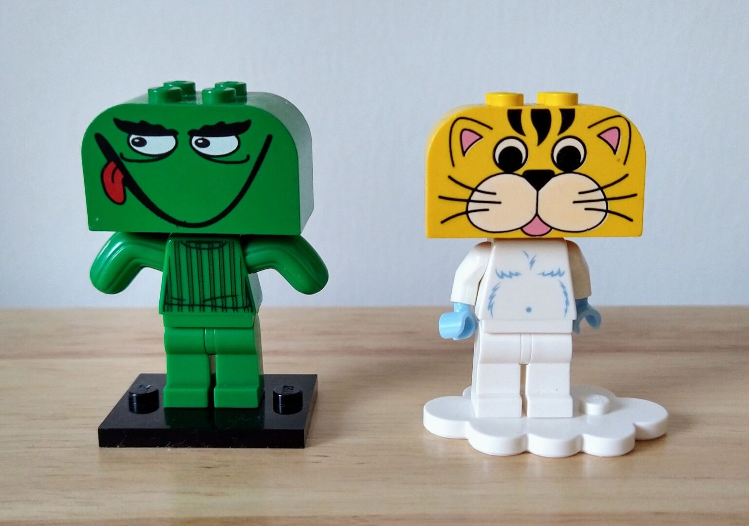 Two Lego minifigures with their heads replaced with a larger 4x2x2 old head pieces. One resembles a cactus with a mischievous face, the other have a white body with a yellow cat head.