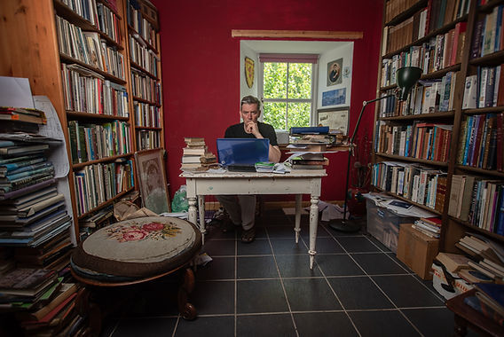 Red room with tall, packed bookcases with a cluttered table behind which John sits looking at the camera.