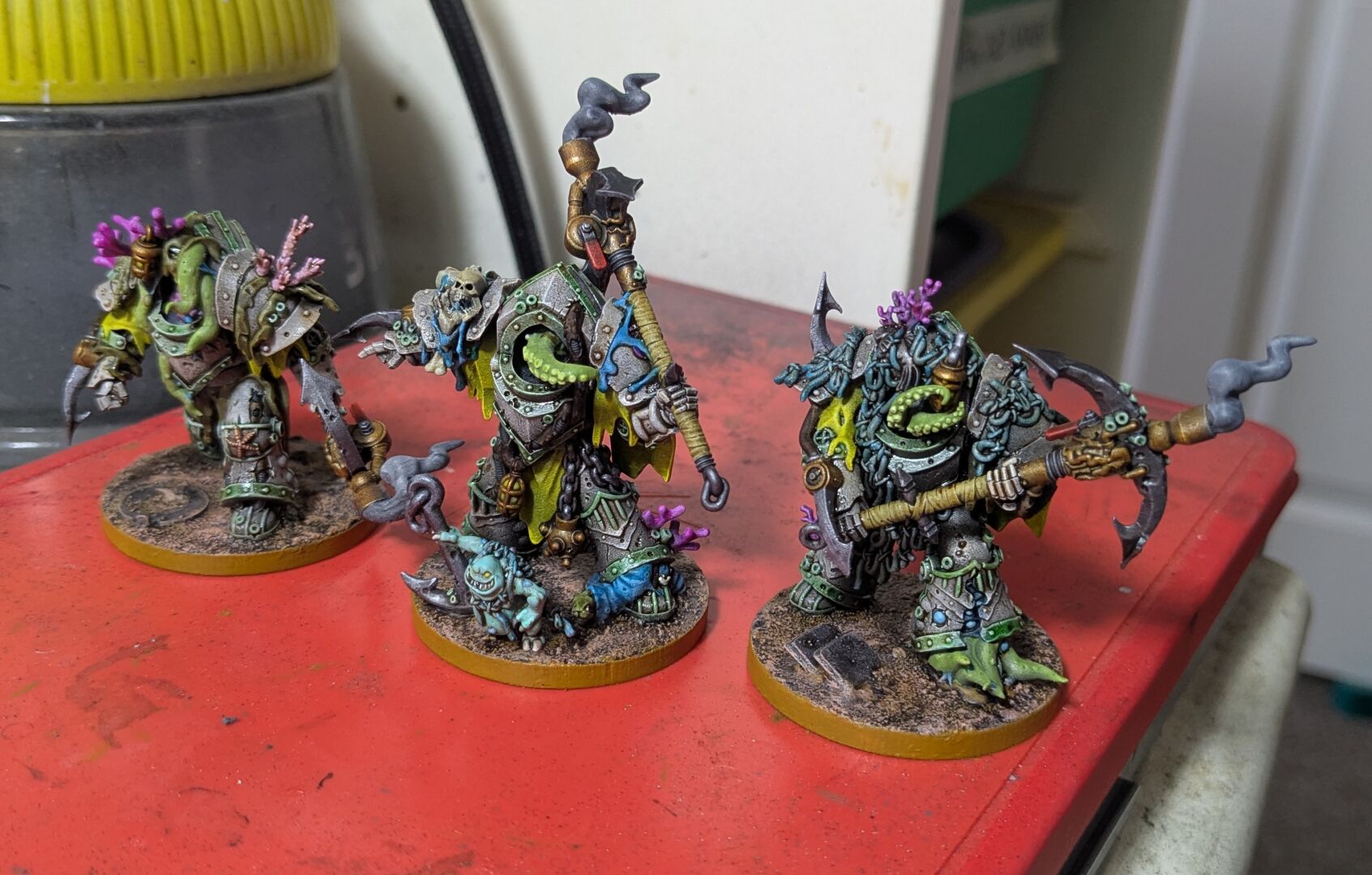 Three painted wargaming miniatures - the Deep Shroud from Mezgike.
They're essentially sci fi humans in powered armour with lots of nautical theming. Tentacles, coral, barnacles, and fishing nets feature heavily.

The miniatures are painted to have white armour with faded green edging. They look like it has been at the bottom of the sea for a long time.