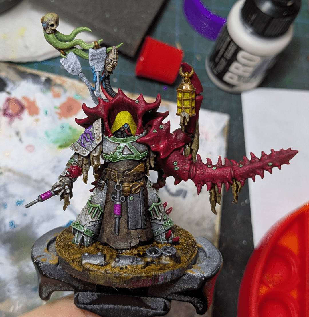 A wargaming miniature on a painting handle. It's nearly done but lacks several steps and so looks unfinished. The miniature is a humanoid in power armour but with one arm replaced by a large crustacean protrusion covered in spikes. Other than that various bits of medical paraphernalia is scattered across him.