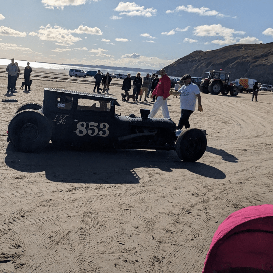 looking along a beach, the headlands visible in the background. In the foreground is a very low, chopped hotrod, possibly based on a car from the 1920s. The car is entirely black and has had its axles moved to beyond the ends of the body. Decorative air vents cover the roof, mirroring the vents on the bonnet.